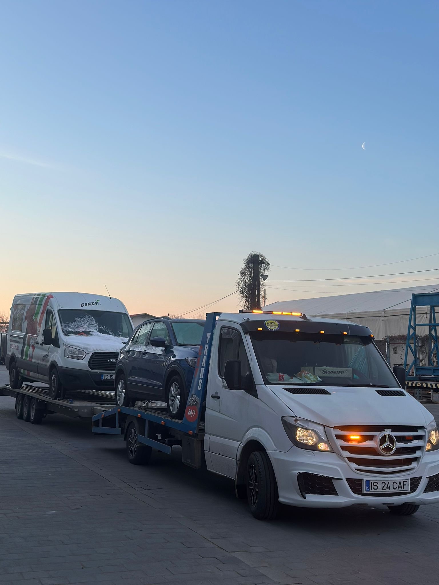 Autoplatforma cu SUV albastru si duba Ford Transit pe remorca, fotografiata la apus